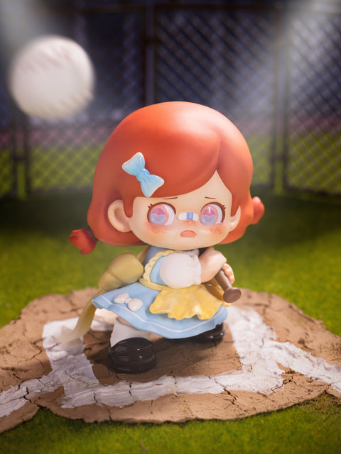 KO Power figure presents a girl with red hair and a blue bow, dressed in a patched dress, standing in a baseball field. She grips a bat nervously as a baseball approaches, with spotlight beams overhead. Her wide-eyed expression and bandages emphasize fragile strength, grit, and determination under pressure.