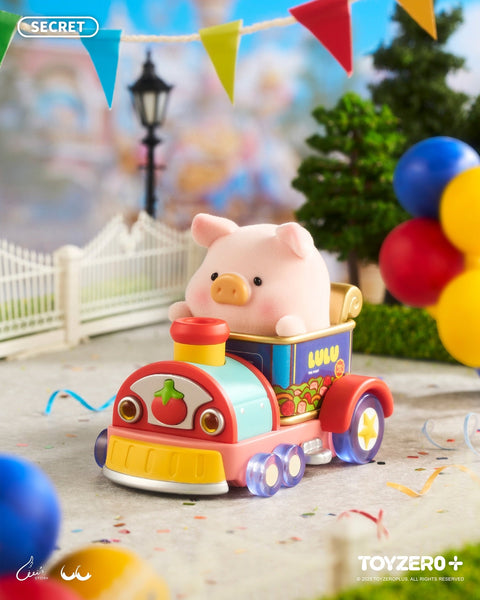 A pig figurine rides in a brightly colored mini train car decorated with fruit graphics. Balloons, trimmed hedges, white fencing, and scattered confetti surround the cheerful park pathway under soft daylight.