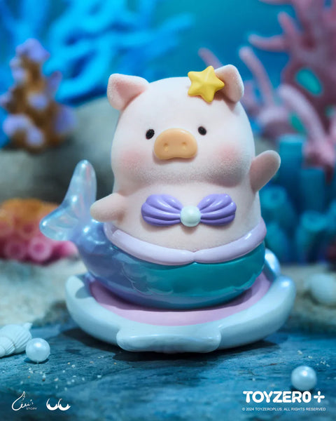 A LuLu the Piggy figure from the Ocean Series by 52TOYS and ToyZero+ appears as a charming mermaid. The pig wears a sparkly pastel purple seashell bra, a shiny blue mermaid tail, and a yellow star on its head. It sits on a shell-like base surrounded by underwater scenery with coral, seashells, and pearls. With a soft flocked texture and serene expression, the figure evokes a magical undersea fairy tale.