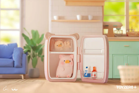 In a pastel-toned kitchen scene from the “Lulu the Piggy: Stay With You” blind box series, a pink piggy figurine is adorably tucked inside a mini pink refrigerator. One pig rests in the freezer compartment while another sits on the main shelf below. The fridge door is open, revealing miniature bottles of milk and sauce. Behind them, a cozy interior with mint green cabinets, a mixer, and a blue couch completes the playful, whimsical setting filled with warmth and charm.