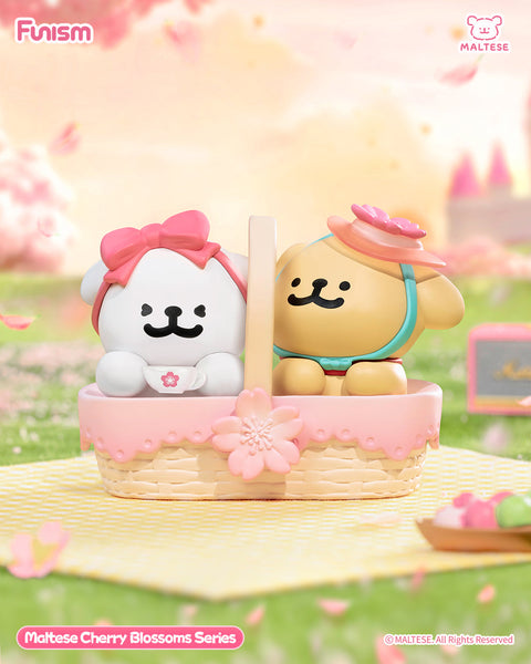 Two Maltese bears sit inside a pink picnic basket, one white with a bow and one tan with a pink flower hat. The basket sits on a pastel mat surrounded by cherry blossoms, evoking a joyful picnic atmosphere.