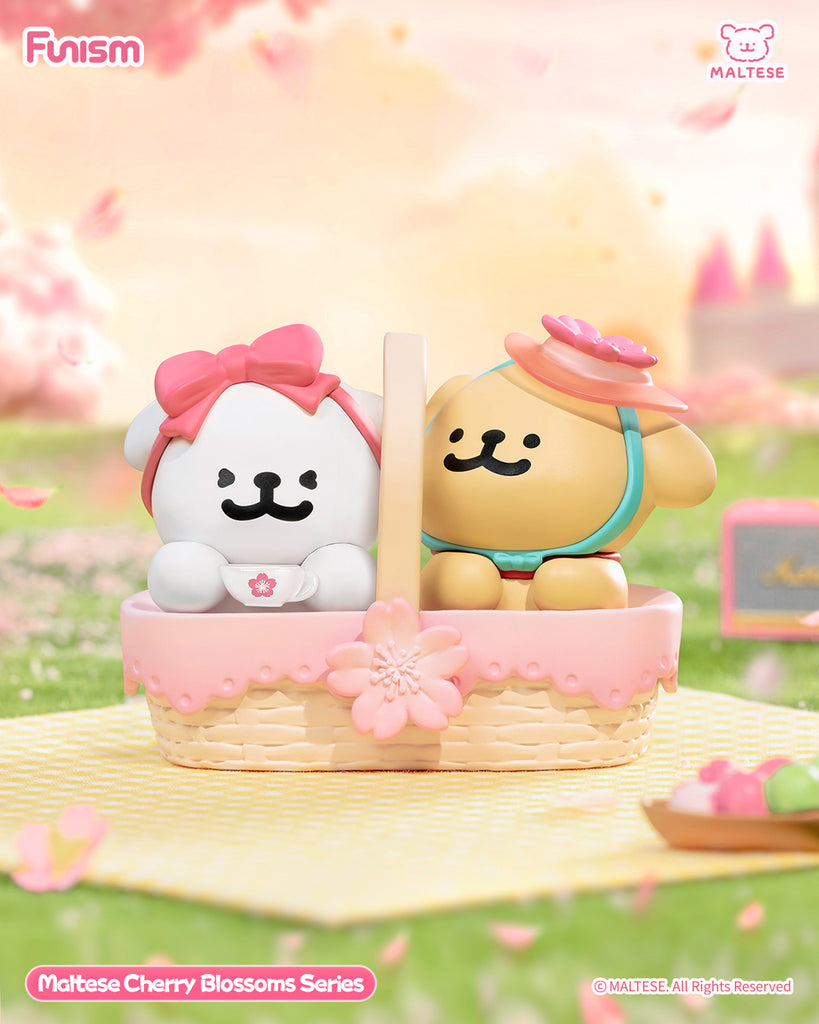 Two Maltese bears sit inside a pink picnic basket, one white with a bow and one tan with a pink flower hat. The basket sits on a pastel mat surrounded by cherry blossoms, evoking a joyful picnic atmosphere.