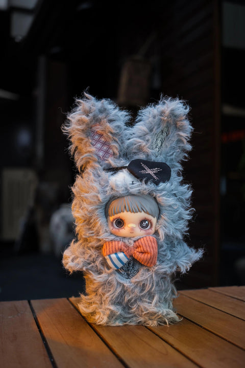 Grey shaggy plush doll with long bunny ears, a black eyepatch with a cross, and a striped orange and blue bow tie. The eyes feature intricate circular patterns.