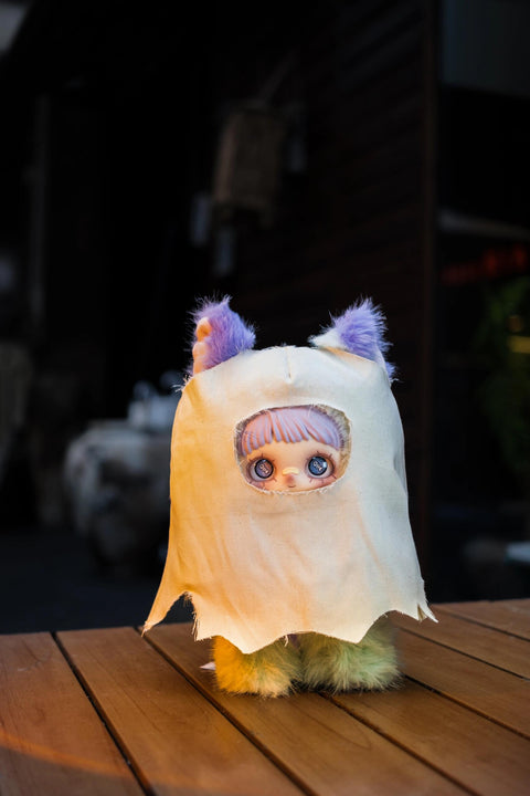 Plush doll wearing a cream-colored ghost sheet with a jagged hem and a cutout for its face. Purple furry ears poke out from the top of the sheet.