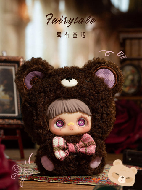 A plush figure sits centered on a patterned rug inside a warmly lit interior scene. The character wears a dark brown fuzzy bear hood with rounded ears and a small cream nose detail on top. A pink and brown plaid bow is tied at the neck. The face shows large purple eyes, short brown bangs, and a neutral expression. Background props include framed art, candles, and soft furnishings.