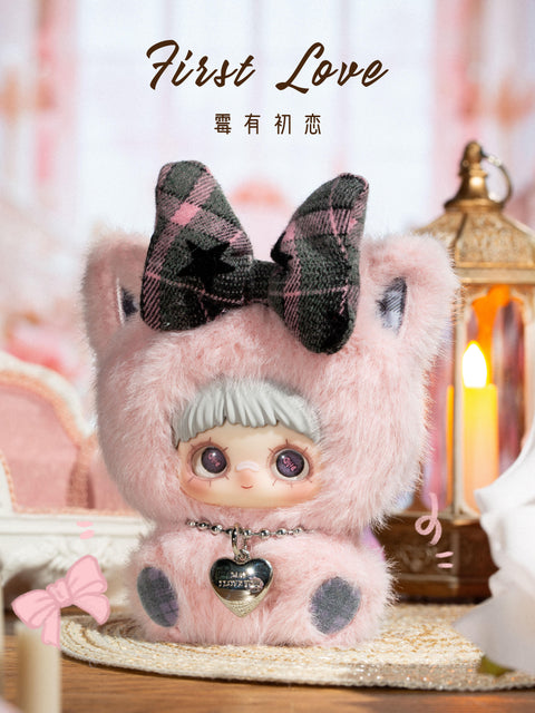 A plush figure stands on a tabletop wearing a pale pink fuzzy bear hood with rounded ears and a large dark plaid bow positioned between the ears. The character has light gray bangs, wide printed eyes, and a calm expression. A silver heart-shaped pendant hangs from a beaded necklace at the chest. The background includes a glowing lantern, soft fabric textures, and warm ambient lighting.