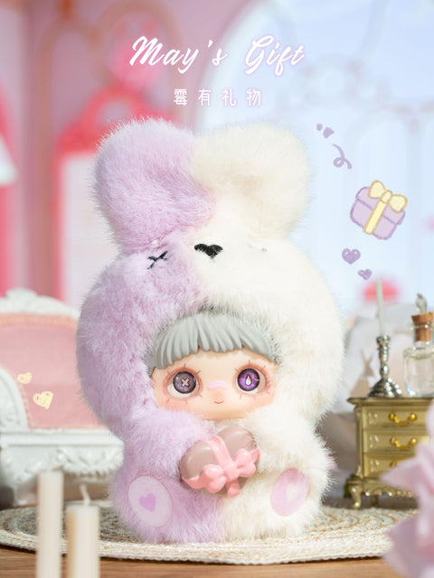 A plush figure is shown holding a translucent pink heart-shaped object tied with a molded bow. The character wears a two-tone fuzzy hood split between pale pink and cream, with rounded ears on top. The face has gray bangs, large eyes with decorative highlights, and a small smile. The figure stands on a rug within a softly lit room featuring pastel furniture and decorative candles.