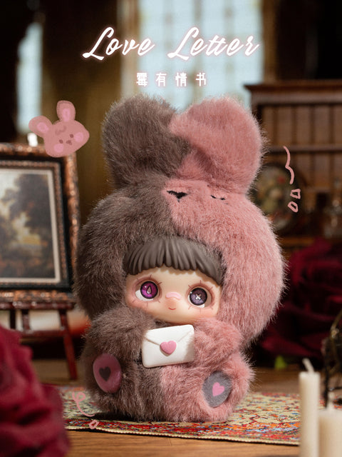 A plush figure holds a small white envelope sealed with a pink heart. The character wears a fuzzy hood split between brown and dusty pink, with rounded ears and stitched facial markings on the hood. The face features brown bangs and large purple eyes. The figure is placed on a patterned rug in a dim, cozy room with framed artwork, candles, and soft furnishings.