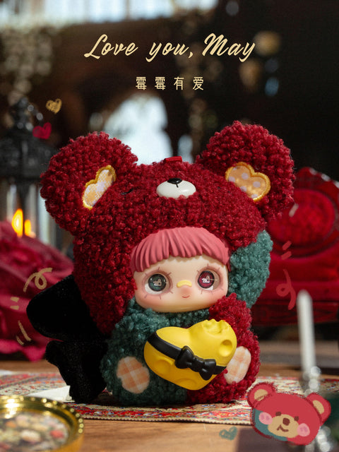 A hidden chaser plush figure is shown wearing a textured red fuzzy bear hood with heart-shaped ear patches and rounded ears. The character has pink bangs and mismatched decorative eyes. A yellow heart-shaped object with a black bow detail is held at the front. The figure sits on a patterned rug with red roses, candles, and dark romantic background elements.
