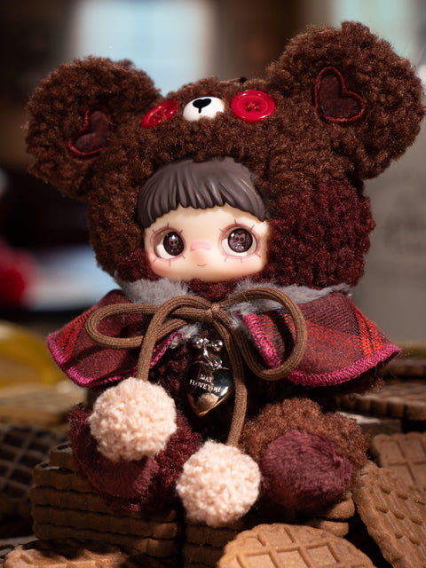 A plush figure dressed in a deep brown fuzzy bear hood sits on stacked cookies. The hood has red button eyes and stitched heart patches on the ears. The figure wears a dark plaid cape tied with brown cords and pom-poms. Warm brown plush textures and dessert props surround the character.