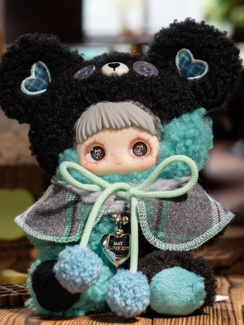 A plush character wears a black fuzzy bear hood with gray button eyes and teal heart-shaped ear patches. The figure has short gray bangs and printed eyes. A gray and green plaid cape is tied with mint pom-pom cords, and the plush body mixes black, teal, and mint textures.