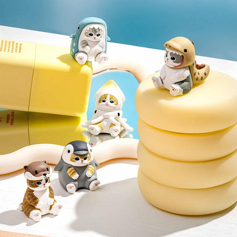Five small cat figurines dressed as various sea creatures sit against a plain white background. One wears an otter hood, another a beige shark hood with teeth framing the face, a third is in a blue spotted whale outfit, a fourth appears as a gray penguin, and the last wears a white squid costume with dangling arms.