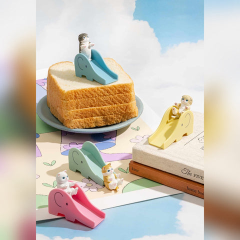 A lifestyle image featuring four miniature cat figures interacting with colorful elephant-shaped playground slides. A grey tabby sits at the top of a blue slide, a cream cat slides down a yellow one, a white cat sits by a pink slide, and a ginger cat is perched on a mint green slide.