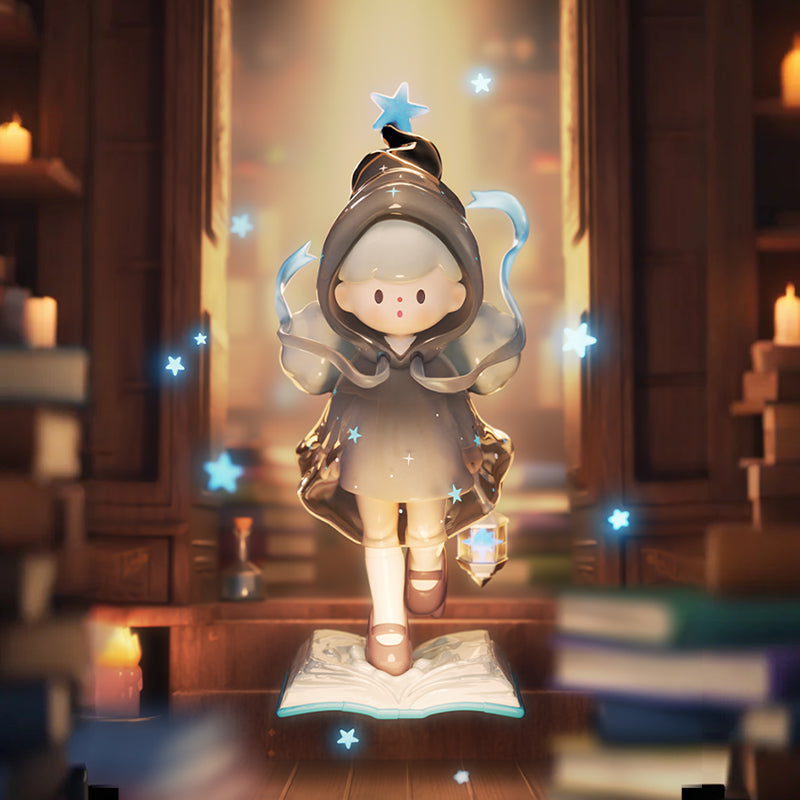 A vinyl figure in a glossy gray hooded cloak with a small star on the tip steps forward on top of an open book base. Translucent ribbon-like curls trail from the hood, a faceted crystal charm hangs at the figure’s side, and tiny blue star lights float around. The background is a warm, blurred library with bookshelves and candlelight.