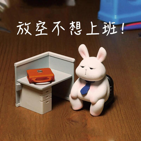 A grumpy white bunny figurine in a blue tie sits slouched at a miniature office desk with an orange briefcase, embodying the Monday blues. The expression shows boredom and reluctance to work.