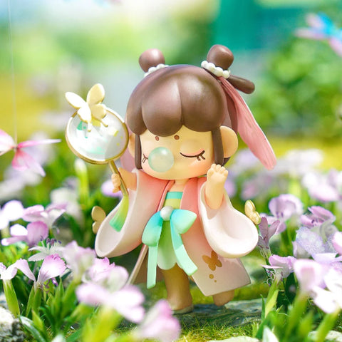 A whimsical collectible figurine from the "Nanci Blooming Girl" series features a girl dressed in a pastel pink and green traditional outfit, walking through a lush flower field. She holds a delicate fan with a butterfly resting on it, her eyes closed in a peaceful expression. Her twin-bun hairstyle is adorned with pearls and a flowing ribbon. The vibrant garden setting, filled with blooming flowers and soft sunlight, enhances the serene and enchanting atmosphere of this tranquil moment.