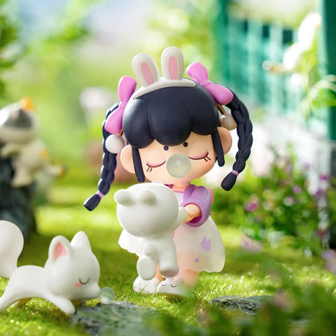A charming collectible figurine from the "Nanci Blooming Girl" series features a girl dressed in a pastel outfit with bunny-themed details, wearing a headband with bunny ears. She playfully swings a small white rabbit in her hands while blowing a bubblegum bubble. Other fluffy animals frolic nearby in the lush green garden. With braided hair tied in pink ribbons and a joyful expression, the scene captures a heartwarming and carefree moment in nature.