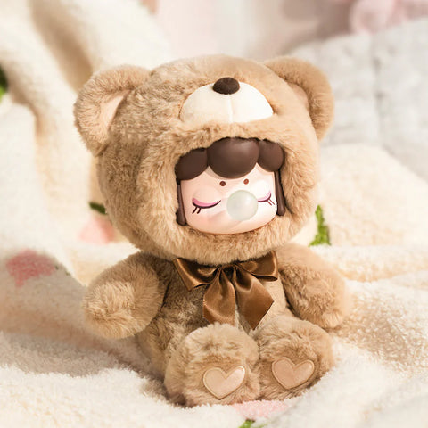 A cozy collectible figurine from the "Nanci Colorful Chocolate Cutie Bears" plush series features a character dressed in a soft, caramel-brown bear suit with a matching satin bow. The figure has a peaceful expression with closed eyes and a bubblegum bubble, while heart-shaped details on the plush paws add extra charm. Seated on a fluffy blanket, the warm lighting and gentle textures create a comforting, snuggly atmosphere, perfect for a dreamy and relaxing moment.