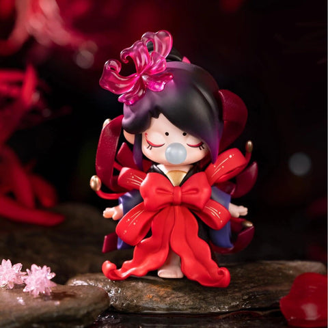 A figurine from the Nanci “Flower Stories” blind box series inspired by the camellia flower. The character wears a deep red and black outfit with a large red bow resembling camellia petals, and her dark hair is styled with a translucent red flower. Eyes closed and blowing a bubble, she stands serenely on a stone surface surrounded by a dark, moody setting, evoking strength, elegance, and timeless beauty.