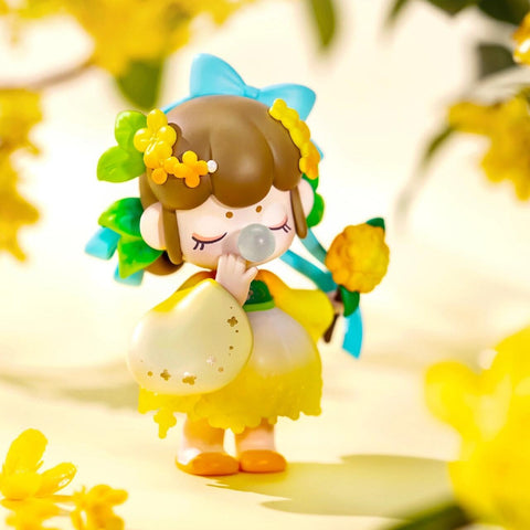 A figurine from the Nanci “Flower Stories” blind box series inspired by the osmanthus flower. The character has short brown hair adorned with yellow blossoms and green leaves, and wears a yellow dress with a petal-like hem and puffy sleeves. Holding a golden osmanthus flower and blowing a bubble, she stands in a sunlit scene filled with soft yellow blooms, capturing the warmth and fragrance of early autumn.