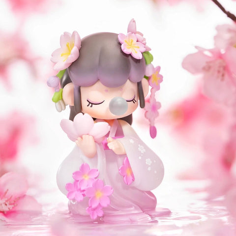 A figurine from the Nanci “Flower Stories” blind box series inspired by the sakura blossom. The character has soft lavender-gray hair adorned with pink cherry blossoms and wears a flowing pink and white hanfu-style dress with floral accents. Eyes closed and blowing a bubble, she holds sakura flowers in her hands while surrounded by floating petals and a dreamy blush-toned setting, capturing the gentle beauty of cherry blossom season.