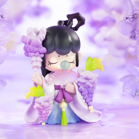 A figurine from the Nanci “Flower Stories” blind box series inspired by the wisteria flower. The character has jet-black hair styled in a unique updo and wears a flowing lavender and blue hanfu-style outfit. Holding a fan and clusters of cascading wisteria blossoms in soft purple and green, she stands serenely with eyes closed and a bubble at her lips, surrounded by dreamy lilac petals that reflect elegance and poetic tranquility.