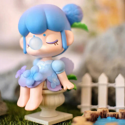 A serene collectible figurine from the "Nanci Secret Garden" series features a character inspired by hydrangeas, dressed in a delicate blue and lavender floral dress. Seated gracefully on a stone pedestal, she has her eyes closed and blows a bubblegum bubble, exuding a peaceful aura. Her hair is styled in a soft blue bun adorned with a hydrangea bloom. Surrounded by a tranquil garden with a pond and white picket fence, the scene captures the essence of a dreamy, everlasting summer.