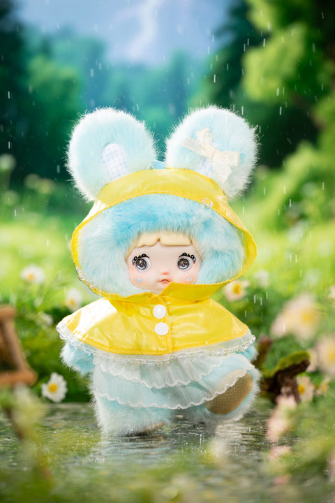 Plush character wearing a pastel blue fuzzy hood with rounded ears and a bright yellow raincoat layered over a frilled dress, posed mid-step in shallow water. Raindrops fall around the figure, highlighting soft textures, translucent trim, and a gentle drizzle weather concept.