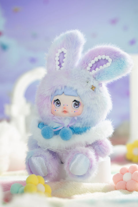 Plush figure seated with a lavender and blue gradient bunny-eared hood, surrounded by a dreamy pastel background. The character features large reflective eyes, tiny star accents on the cheeks, and layered fuzzy fabrics that create a soft, misty atmosphere representing foggy weather.