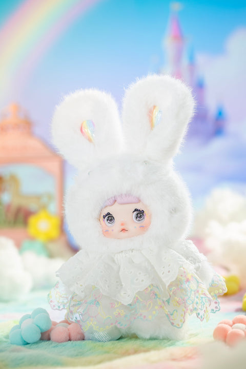 Plush character dressed in a white bunny-eared hood and embroidered pastel skirt, standing against a bright fantasy backdrop with a rainbow and soft clouds. The doll’s glossy eyes, delicate lace details, and airy textures evoke a calm, clear sky after rainfall.
