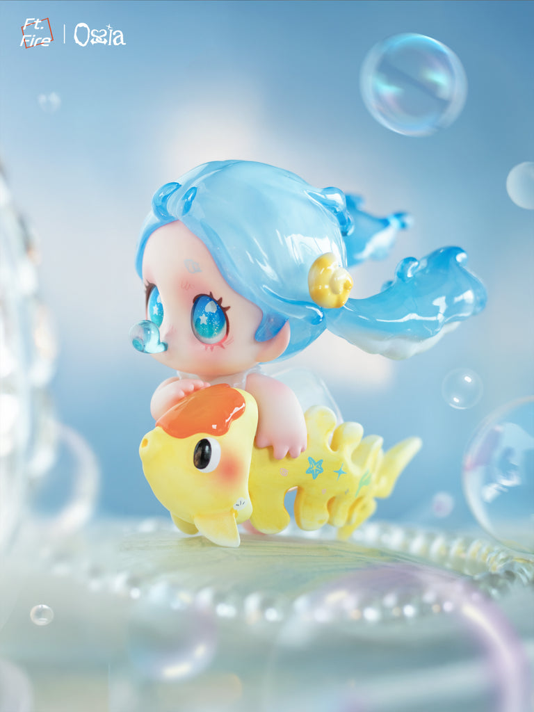 A small vinyl figure with translucent blue hair styled in flowing twin tails rides a yellow fish with an orange head. The character has star-filled eyes and a seashell in her hair, surrounded by floating bubbles in a bright underwater-inspired scene.