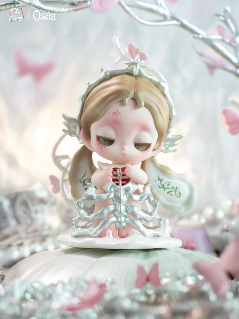 A detailed figurine of a pale angelic character with soft brown hair and sleepy eyes sits on a white base surrounded by silver branches and pink butterflies. The figure holds a silver ribcage that reveals red detailing inside, with sculpted white wings marked by green vine patterns behind her.