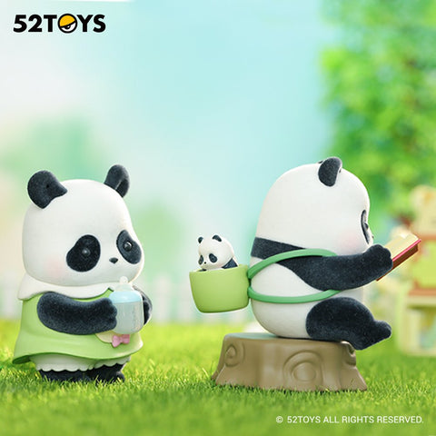 Two panda figurines stand on bright green grass in a soft outdoor scene. One panda holds a baby bottle with both hands while wearing a light green outfit. Nearby, a larger panda sits on a tree-stump seat reading a red book, with a tiny panda peeking from a green pouch on its back.