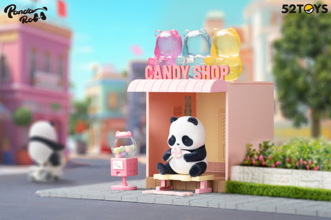 A panda figurine from the 52TOYS Panda Roll Shopping Street series sits on a bench in front of a pastel pink candy shop, holding a pink-and-white lollipop. The shop has a pink awning, a gumball machine, and colorful bear-shaped candy sculptures on the roof. A small candy wrapper lies at the panda’s feet. The background features colorful buildings and a tree-lined street, creating a cheerful, sweet-themed scene in a whimsical miniature town.