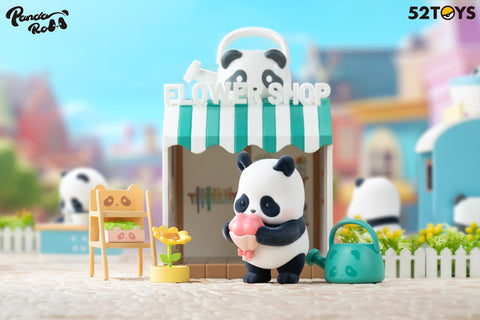 A panda figurine from the 52TOYS Panda Roll Shopping Street series stands outside a charming flower shop with a green-and-white striped awning and a panda-faced watering can on the roof. The panda holds a pink bouquet and is surrounded by garden-themed props, including a sunflower, a small shelf with plants, and a green watering can. The shop’s backdrop includes soft, colorful buildings and a white picket fence, creating a cheerful and inviting atmosphere in this miniature street scene.