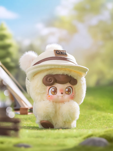 A soft yellow Qkid plush figure stands on green grass wearing a beige explorer hat with the “Qkid” patch on front. The fluffy toy has round eyes with orange star reflections and blush cheeks. The background shows a tent and picnic items in bright morning light.
