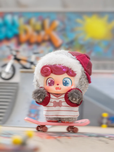 A gray and red plush Qkid figure rides a pink skateboard in front of a graffiti wall and blurred BMX bike. The character has curly magenta hair, mismatched blue and pink eyes, and wears a striped hoodie with a beanie. The scene captures an urban skate park setting.
