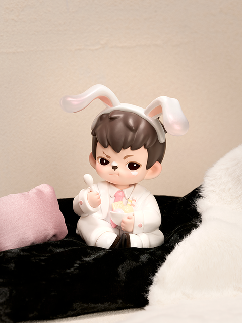 A figure with short brown hair sits on a soft black blanket wearing a white suit and pink tie. Bunny ears rest on its head as it holds a white bowl and spoon, with a small pink pillow nearby, in a cozy beige and white room.