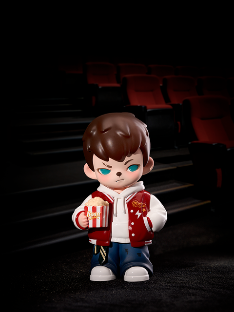 A figure in a red varsity jacket and jeans stands inside a dim movie theater, holding a striped popcorn bucket labeled “Boom.” The red seats and dark shadows create a cinematic atmosphere around the collectible figure.