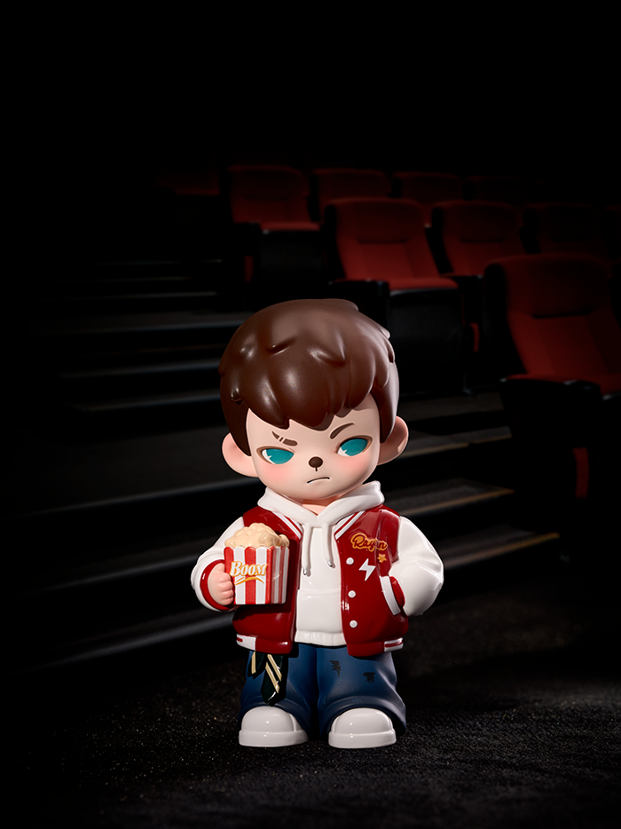 A figure in a red varsity jacket and jeans stands inside a dim movie theater, holding a striped popcorn bucket labeled “Boom.” The red seats and dark shadows create a cinematic atmosphere around the collectible figure.