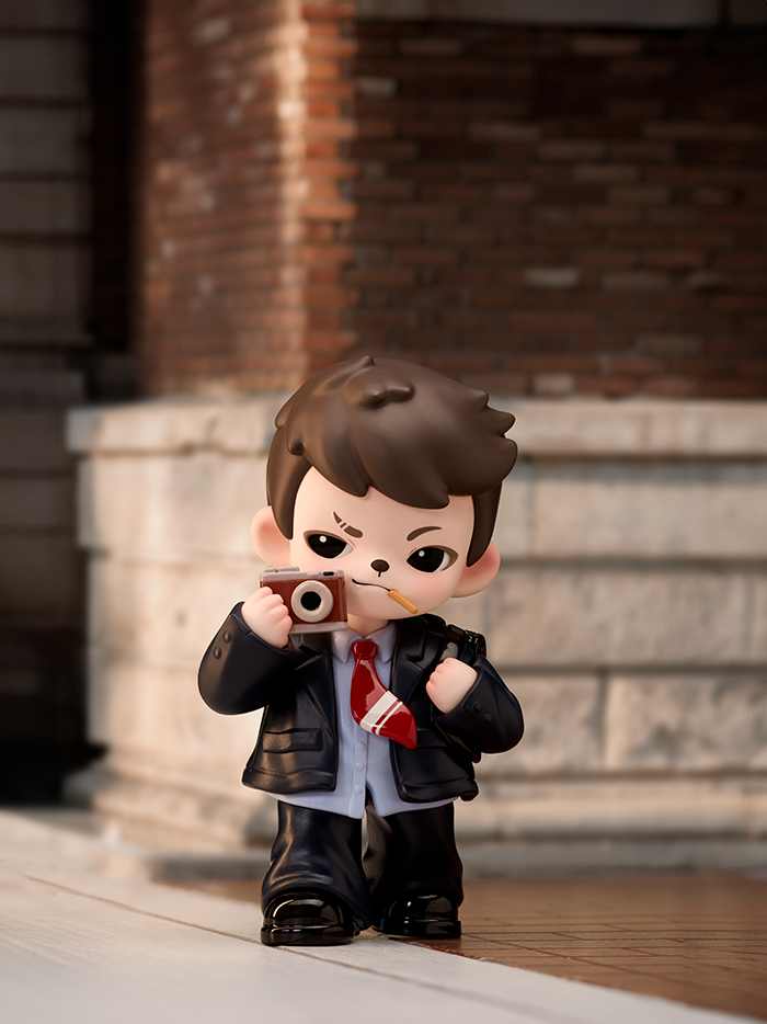 A brown-haired figure in a black suit and red tie holds a camera while chewing a pencil. The figure stands in front of a brick wall background, posing mid-shot with confident energy.