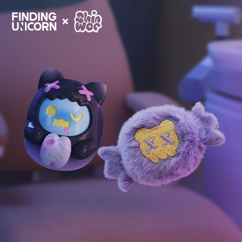 A pair of collectible figures shown in a dim blue room. The left toy is a fuzzy purple candy-shaped plush with a yellow bear face marked with stitched X eyes. The right toy is a glossy black vinyl cat character with pink X marks on its hood, blue face, and yellow eyelids, holding a small pale purple candy.
