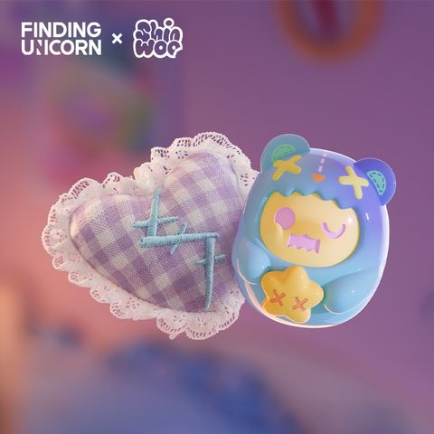 A small blue-and-yellow bear figure floats beside a checkered purple heart-shaped plush. The vinyl figure wears a bear hood with stitched X marks, one closed pink eye, and holds a yellow star with red X’s. The heart plush is edged in white lace and embroidered with teal stitched letters.