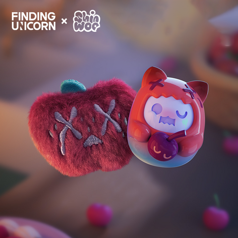 Two matching collectibles shown in a softly blurred kitchen setting. On the left is a fuzzy red apple plush with gray stitched X eyes and a green stem. On the right is a rounded red vinyl cat figure wearing a hood with red X marks and holding a purple cherry with a smiling face.