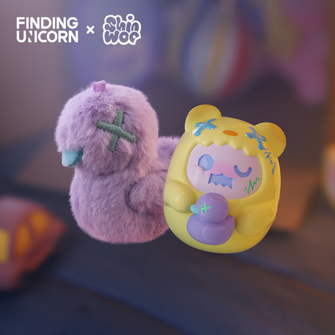 A fuzzy purple plush duck with a blue stitched X for an eye floats beside a yellow vinyl bear figure. The bear wears a hood with blue embroidered markings and holds a small purple duck. Both toys are softly lit against a blurred background with warm highlights.