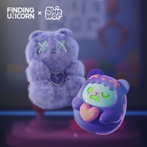 A soft lavender plush bear with stitched teal X eyes and a small embroidered heart on its chest stands beside a glossy purple bear vinyl figure. The vinyl character has green eyes, pink X marks on its hood, and holds a pastel heart reading “HUG ME” in green letters.