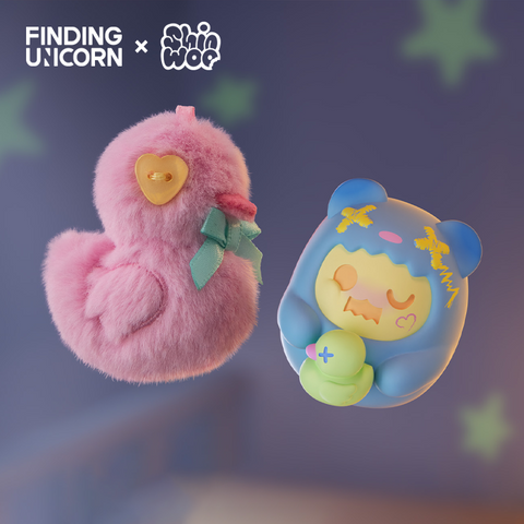 A fluffy pink duck plush with a yellow button heart and green ribbon bow floats beside a blue vinyl bear character. The bear has yellow and pink facial details, wears a blue hood with yellow stitching, and holds a small green duck figure with a stitched X mark.