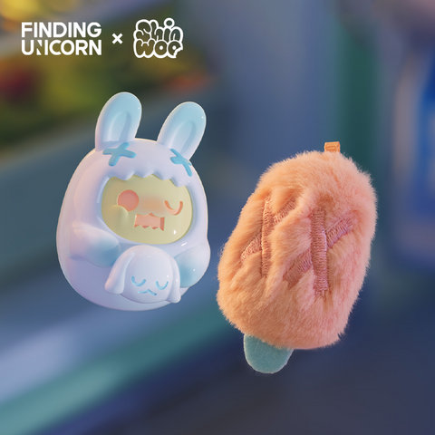 A pale blue vinyl bunny figure with white X marks on its hood floats beside an orange fuzzy popsicle-shaped plush. The plush has stitched pink X marks and a teal handle. The vinyl figure holds a small sleeping bunny toy and glows under soft cool lighting.