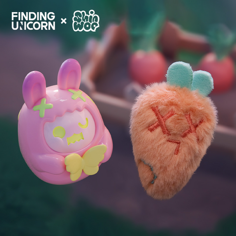 A glossy pink vinyl bunny figure with lime green X marks and a yellow butterfly shape in its arms floats beside a fuzzy orange strawberry-shaped plush. The plush has embroidered red X eyes, a sad stitched mouth, and green leaves on top, set against a blurred garden background.