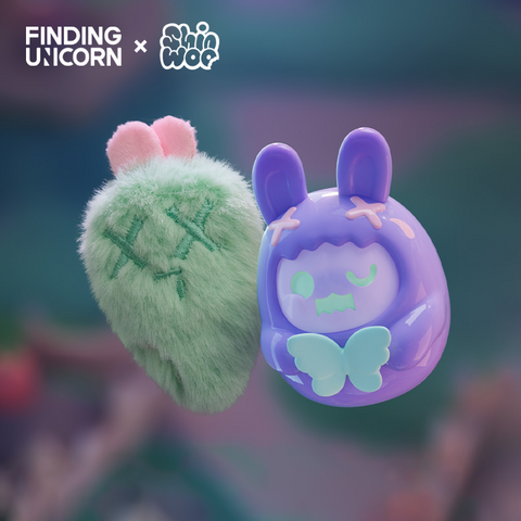 A purple vinyl bunny figure with pink ears and green butterfly details floats beside a fuzzy green strawberry-shaped plush. The plush features stitched X eyes and a small sad mouth. Both figures are softly lit with a blurred greenish background.